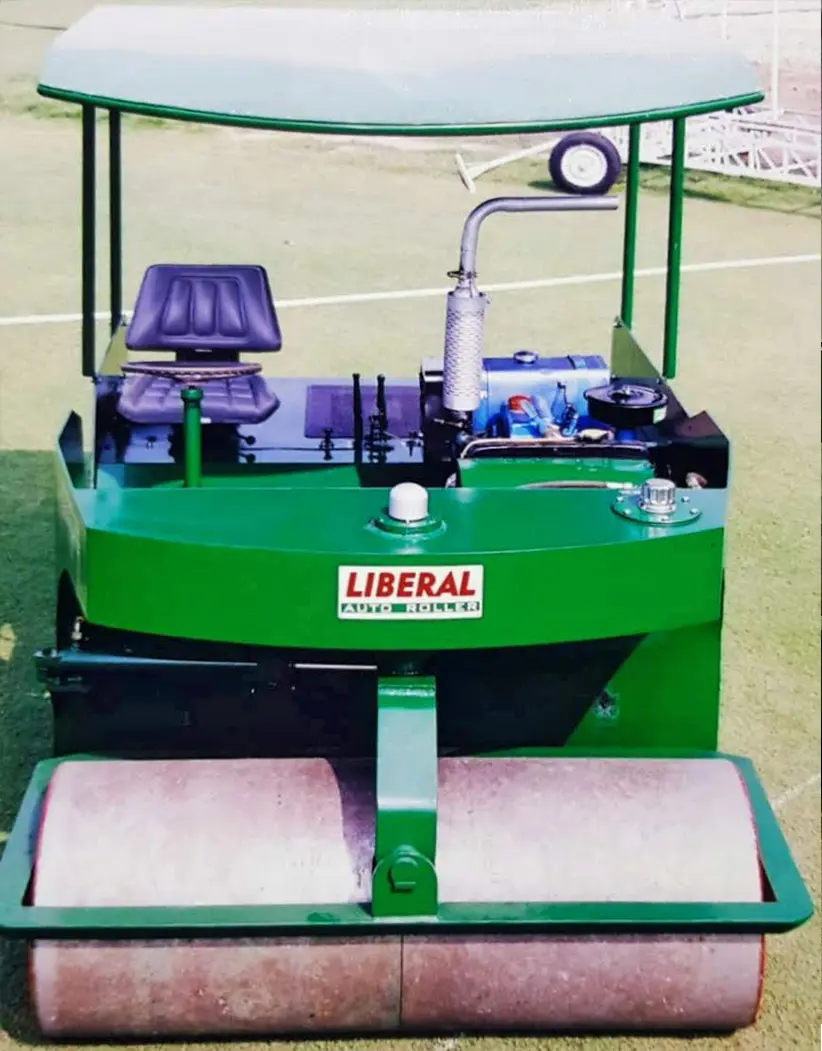 A green and yellow Liberal automatic cricket roller is parked or in use on a pristine cricket pitch at the Kuwait Cricket Club, marking the company's first international machinery export.
